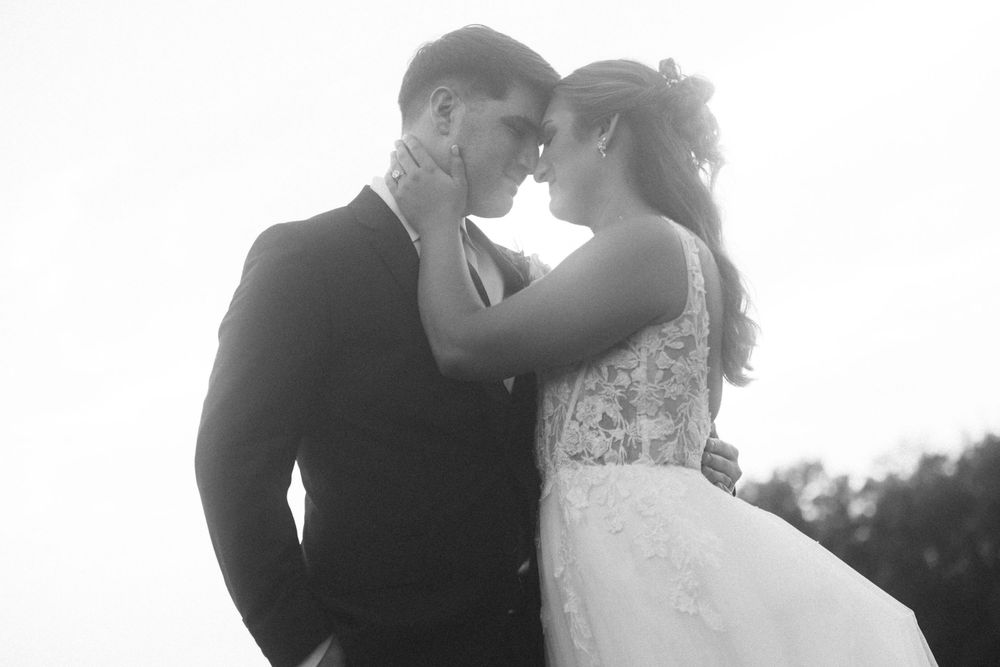 Black and white wedding photo of bride and groom touching foreheads romantically outdoors.