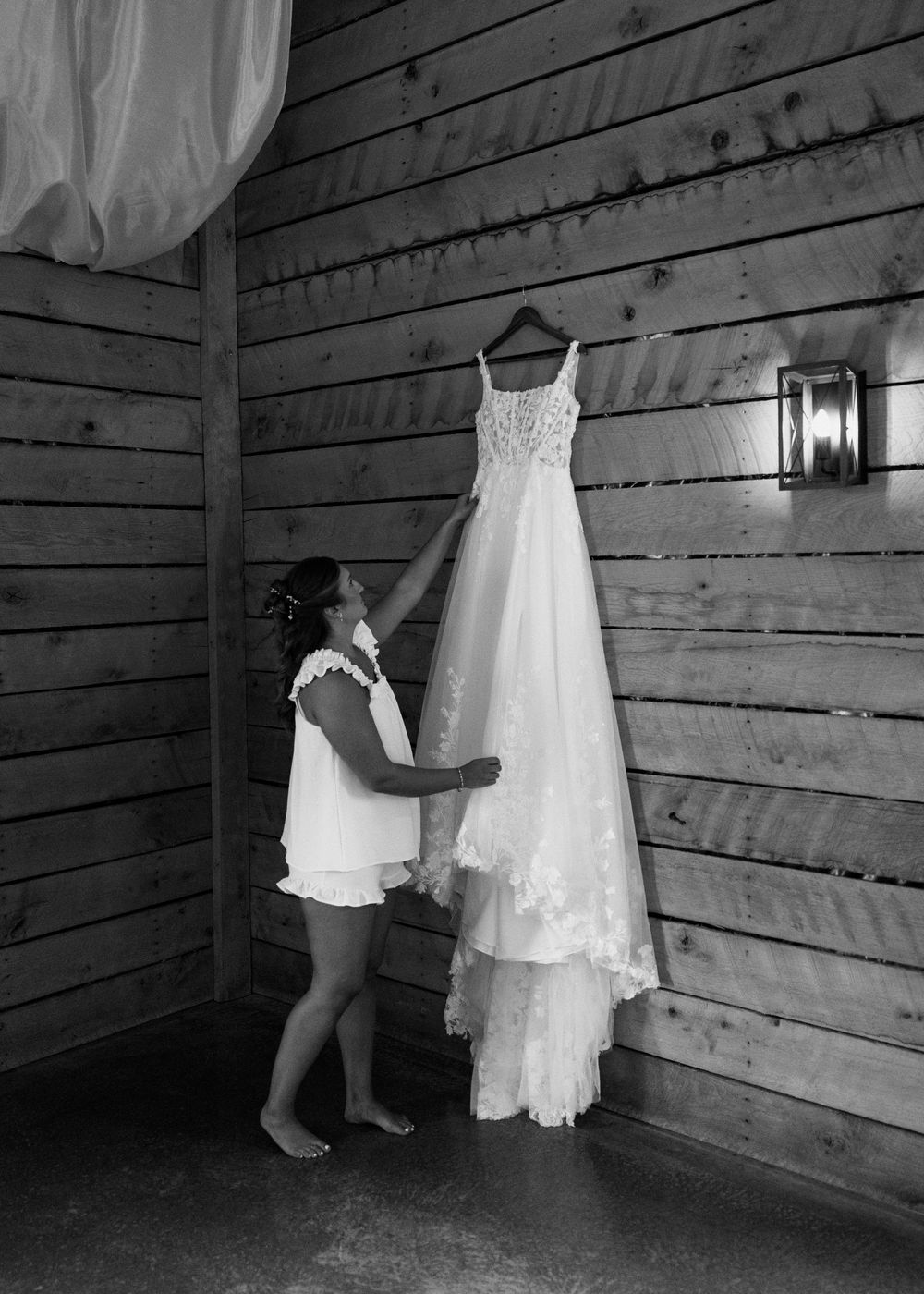 Bride touching her lace wedding gown hanging on rustic wood wall in black and white photo.
