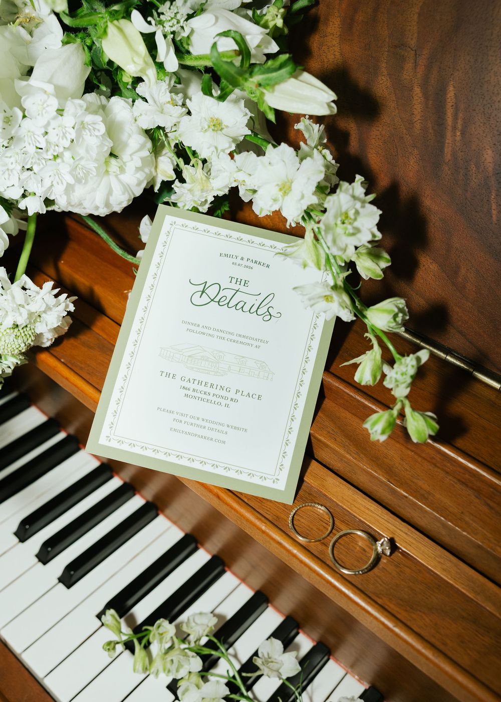 Elegant wedding details card with gold rings and white floral arrangement on a wooden piano.