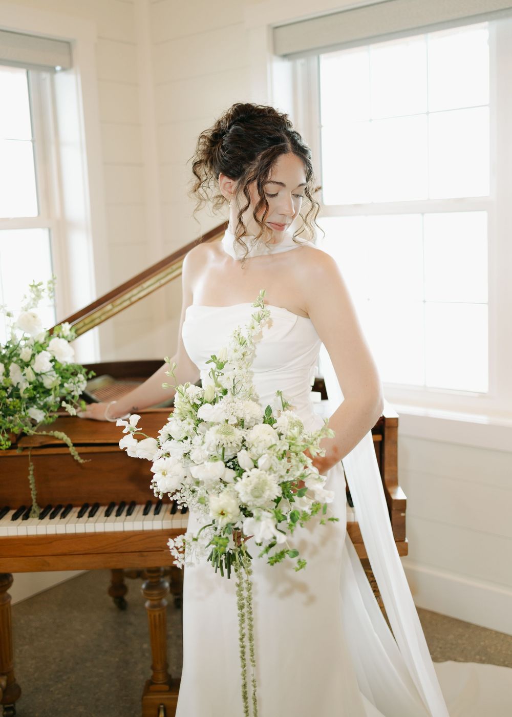 Bride in strapless white gown holding cascading white bouquet beside a vintage grand piano.