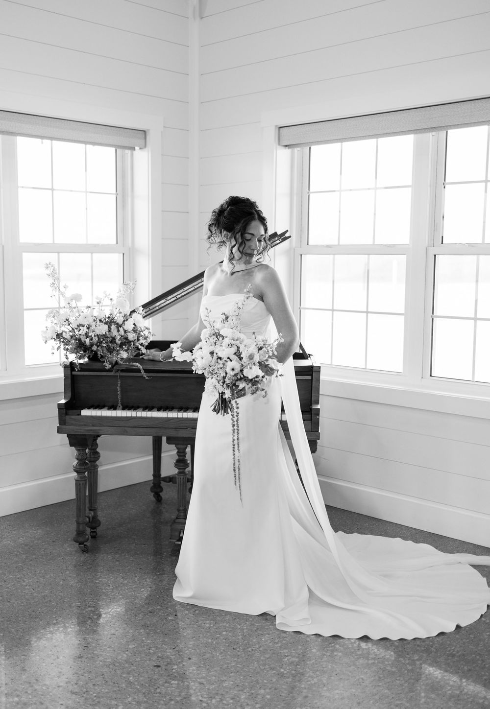 Black and white bridal portrait with bride holding bouquet standing beside grand piano.