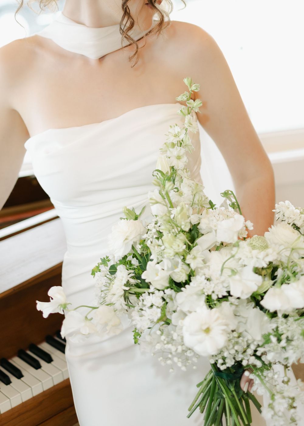 Close-up of bride in strapless white dress holding lush white and green bouquet near piano.