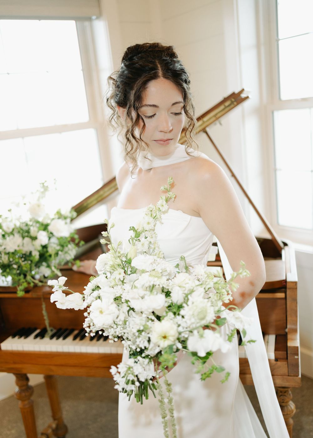 Bride with curly updo in strapless gown holding white floral bouquet beside a grand piano.