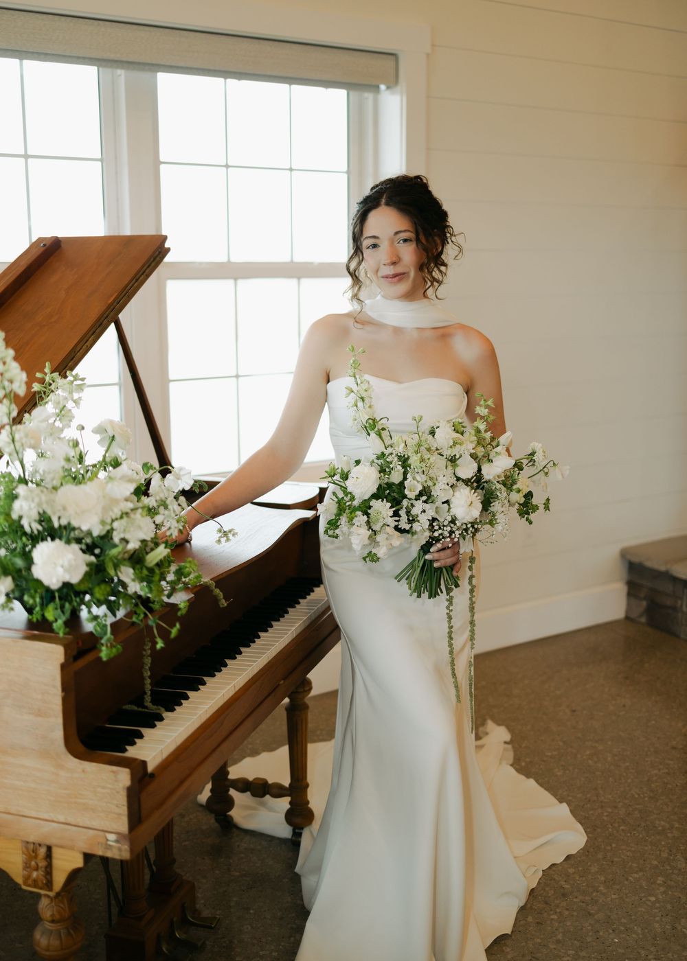 Smiling bride in fitted white gown posing beside a grand piano with white floral arrangements.