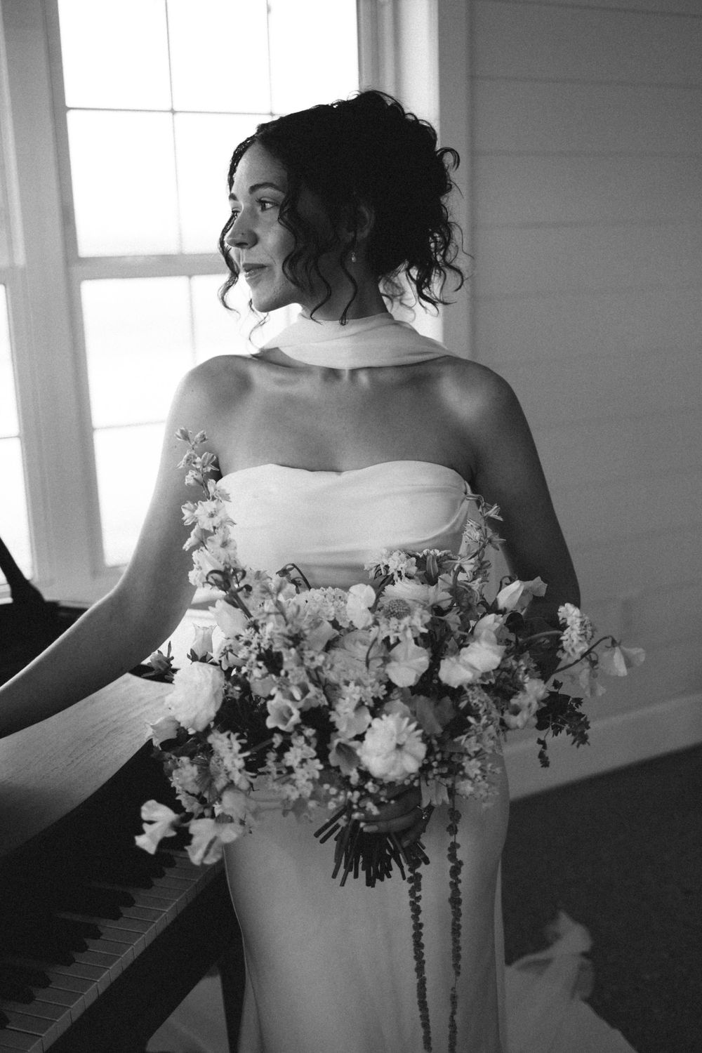 Black and white portrait of bride in strapless gown holding a lush bouquet near a piano.
