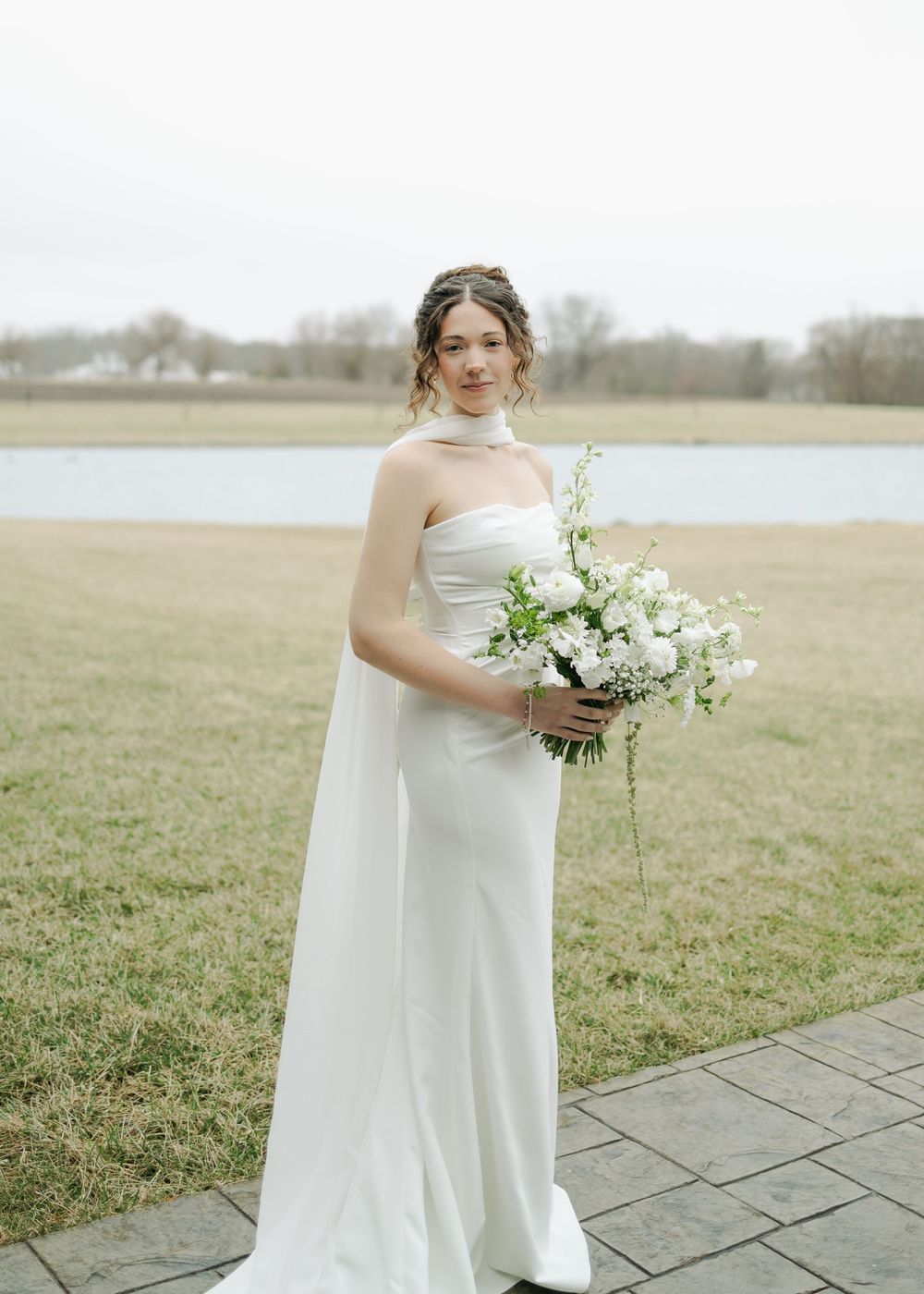 Bride in elegant white gown with scarf holding white bouquet outdoors near a serene lake.