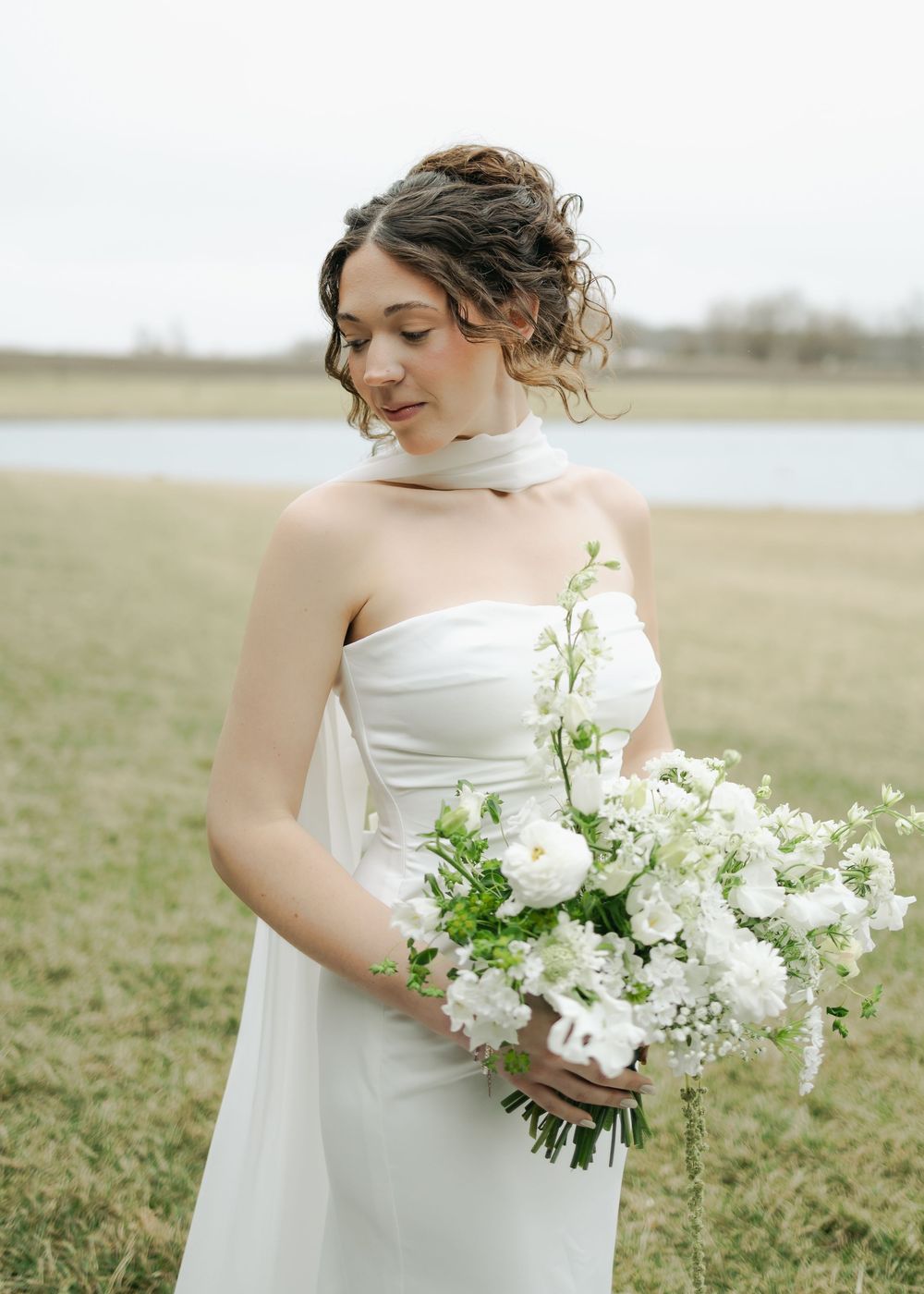 Bride with curly updo in strapless white gown holding white and green bouquet by a lake.
