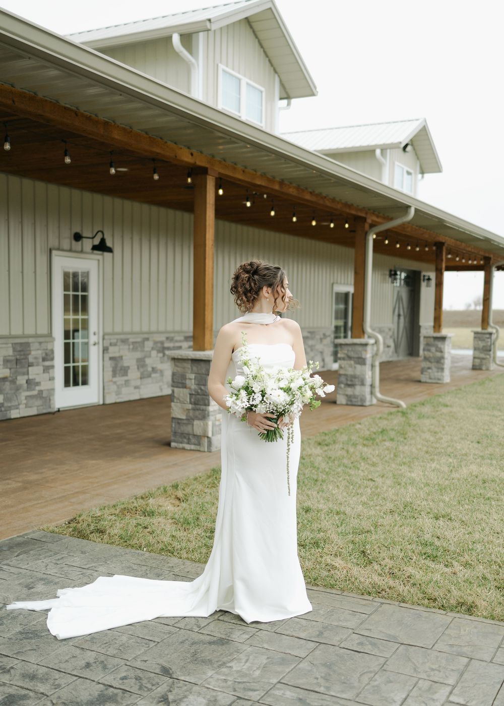 Bride in fitted white gown with train standing outside a modern barn wedding venue.