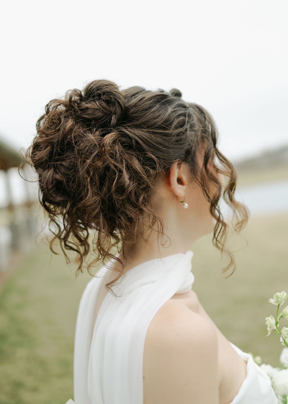 Bride with curly updo hairstyle and pearl earring in white halter wedding dress outdoors.