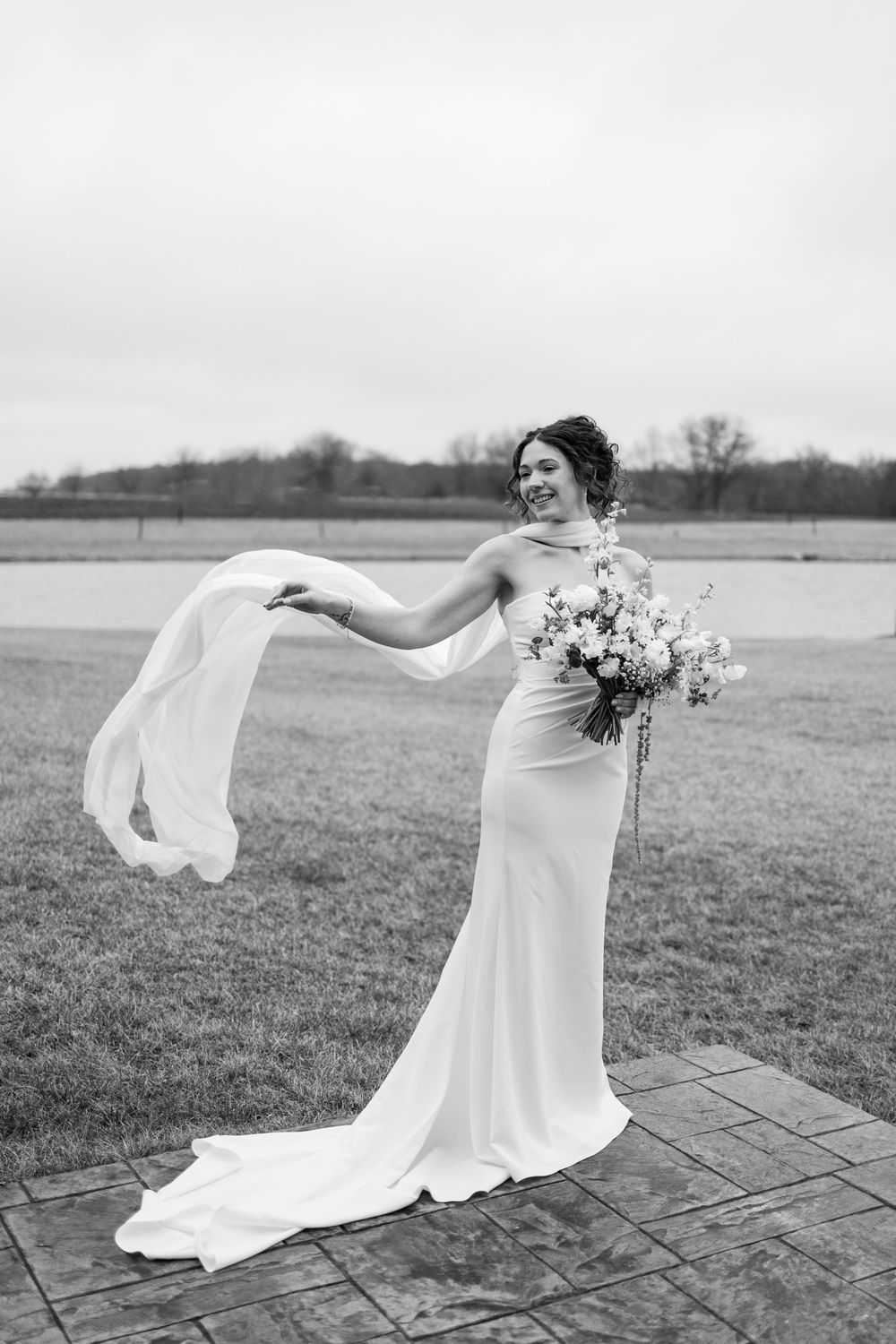 Black and white photo of smiling bride in fitted gown with flowing scarf and bouquet.