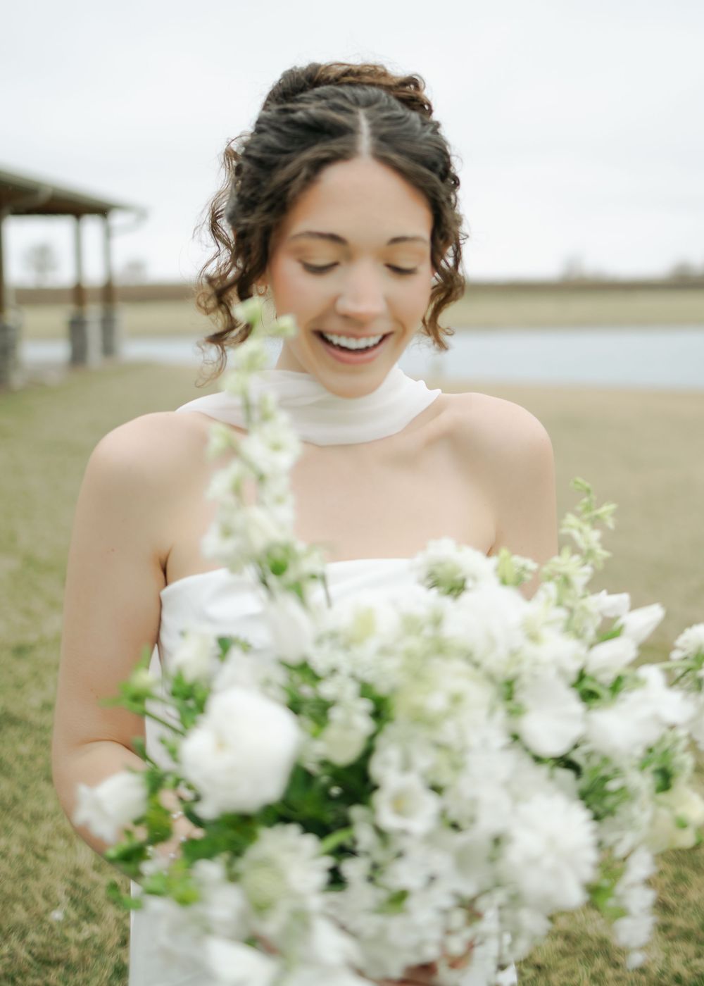 Smiling bride in white halter gown looking down at white and green floral bouquet outdoors.
