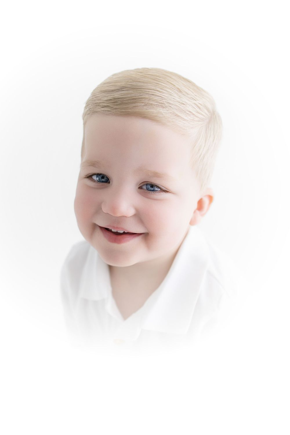 Smiling blonde toddler boy with blue eyes wearing a white shirt against a white background.