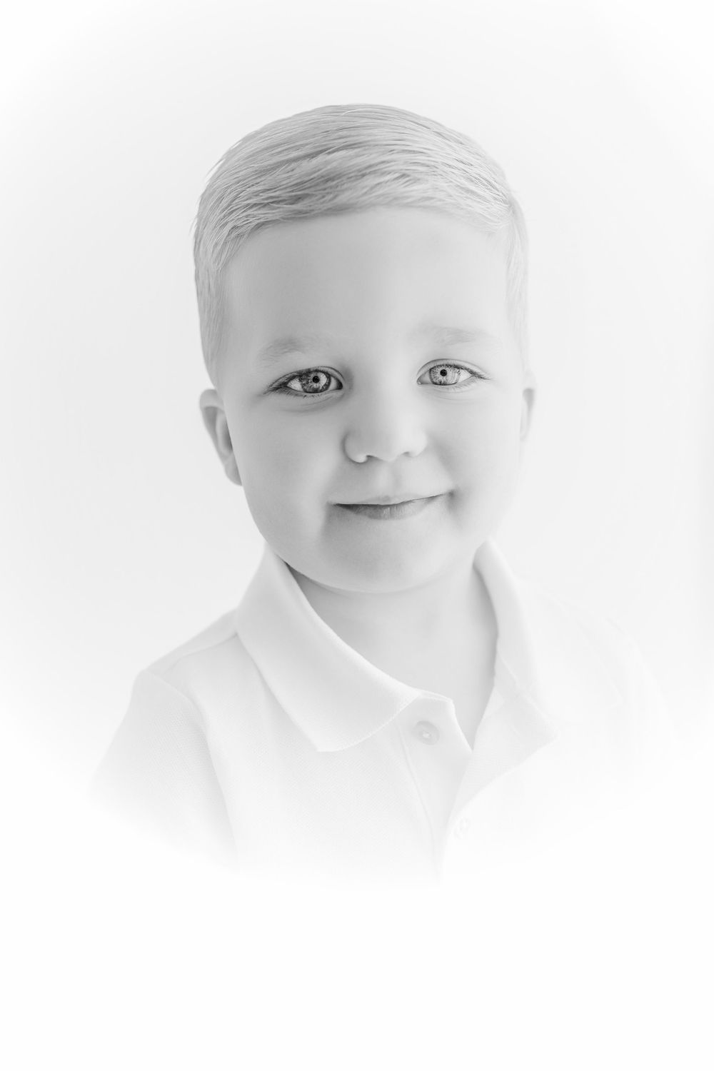 Black and white portrait of a young blonde boy smiling softly, wearing a white polo shirt against a bright background.