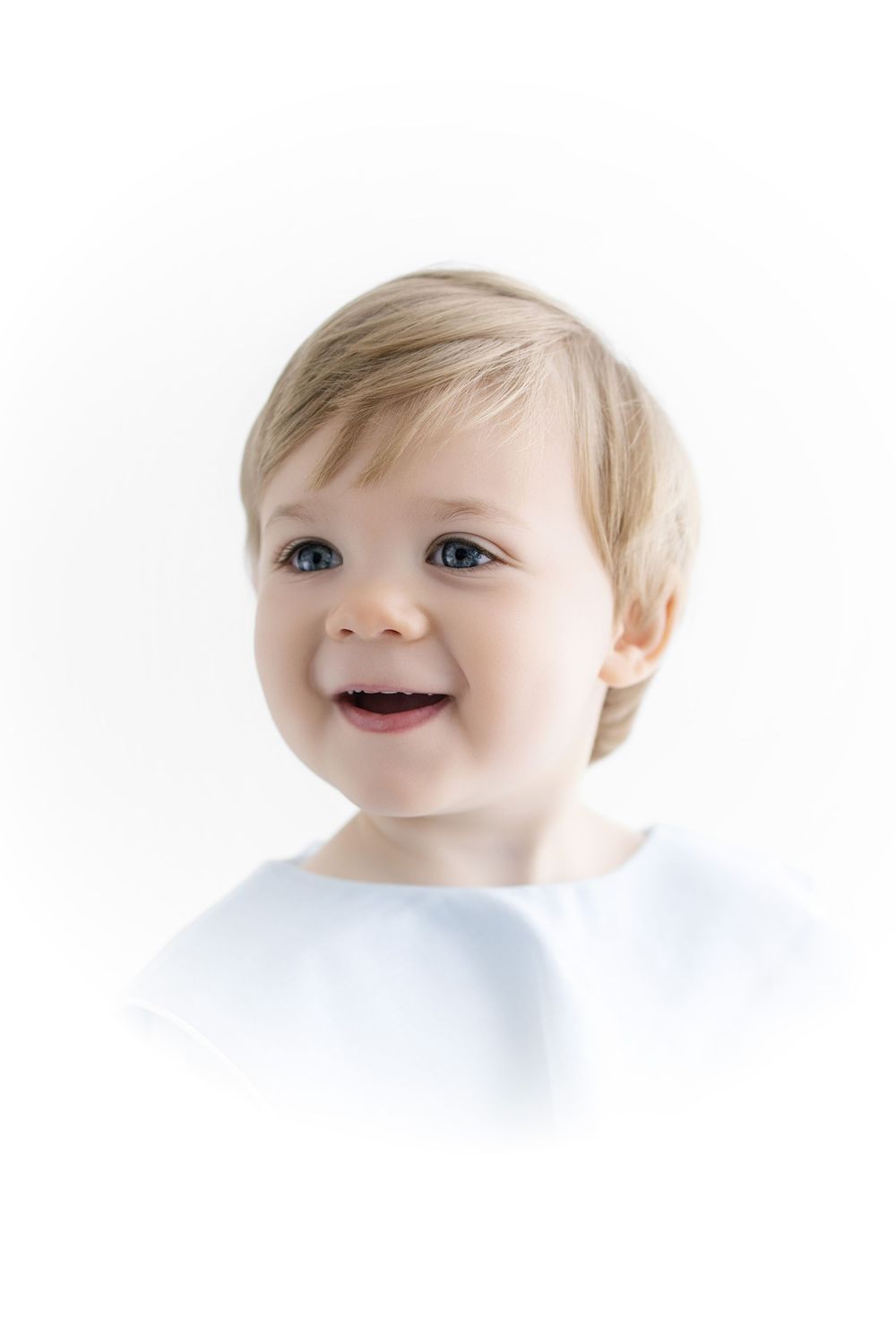 Smiling baby with blue eyes and blonde hair wearing a white shirt against a white background.