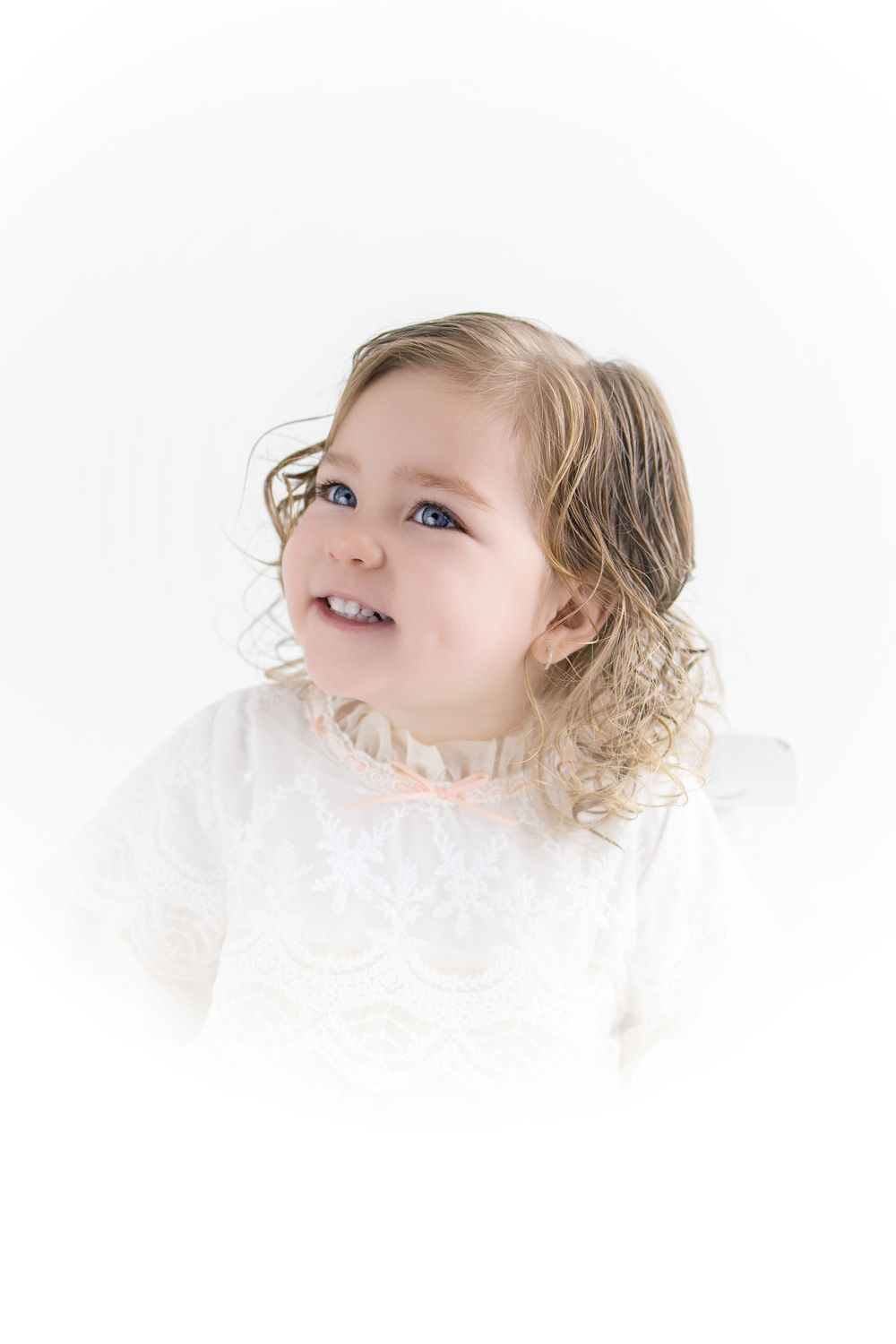 A smiling toddler girl with curly blonde hair and blue eyes wearing a white lace dress on a white background.