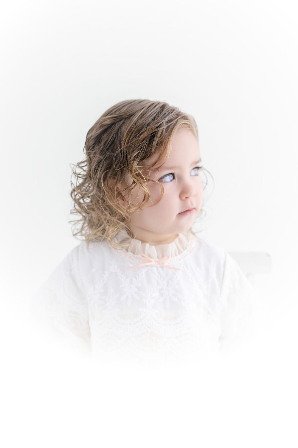 Adorable toddler girl with curly blonde hair and blue eyes wearing a white lace dress against a white background.