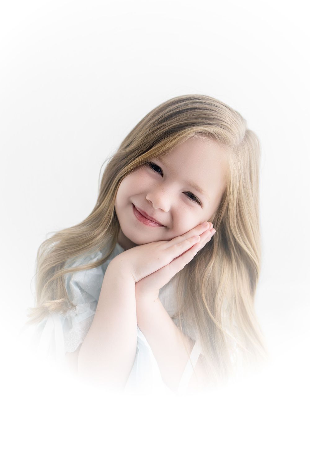 A smiling young girl with long blonde hair rests her cheeks on her hands against a soft white background.