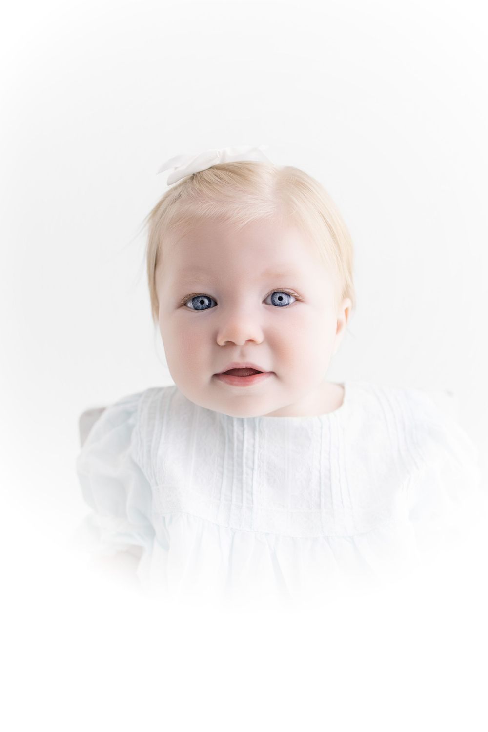 Adorable baby girl with blue eyes and blonde hair wearing a white dress with bow headband.
