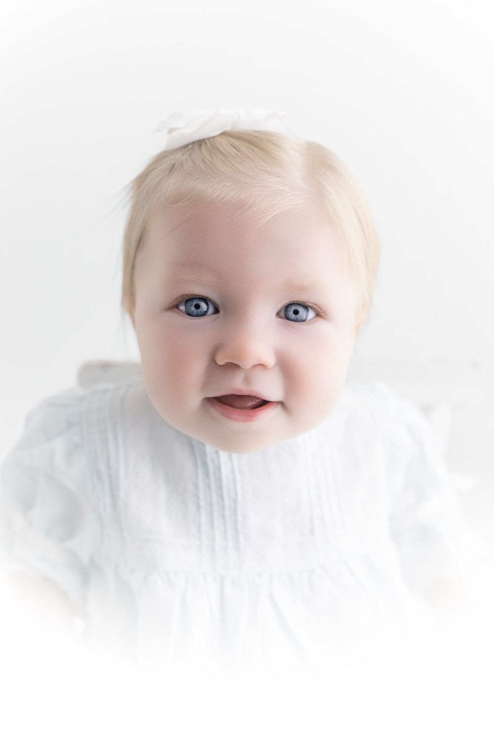 Adorable baby girl with blue eyes and blonde hair wearing a white dress and bow headband on white background.