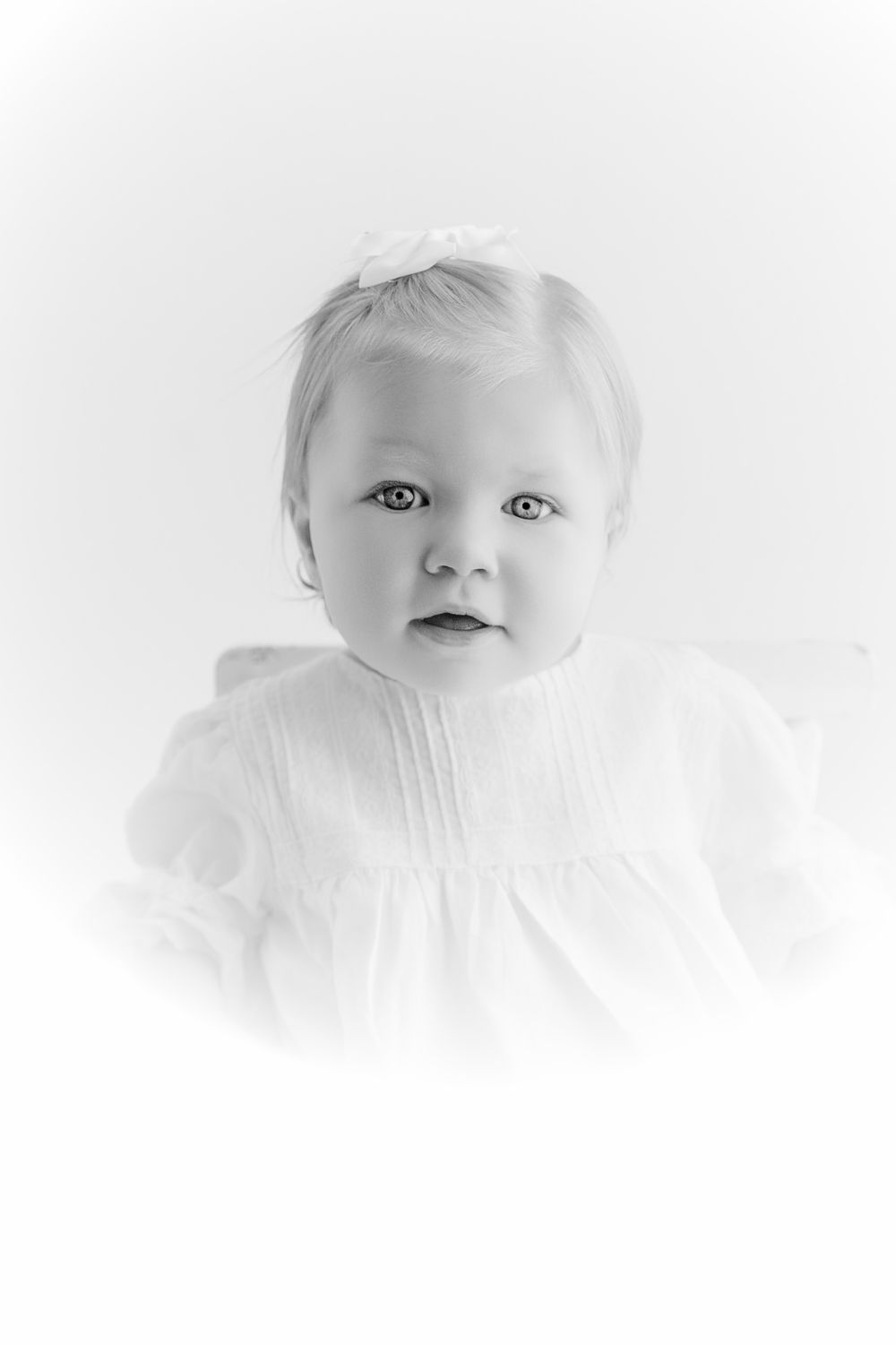 Black and white portrait of a baby girl in white dress with bow, soft vignette studio photography.