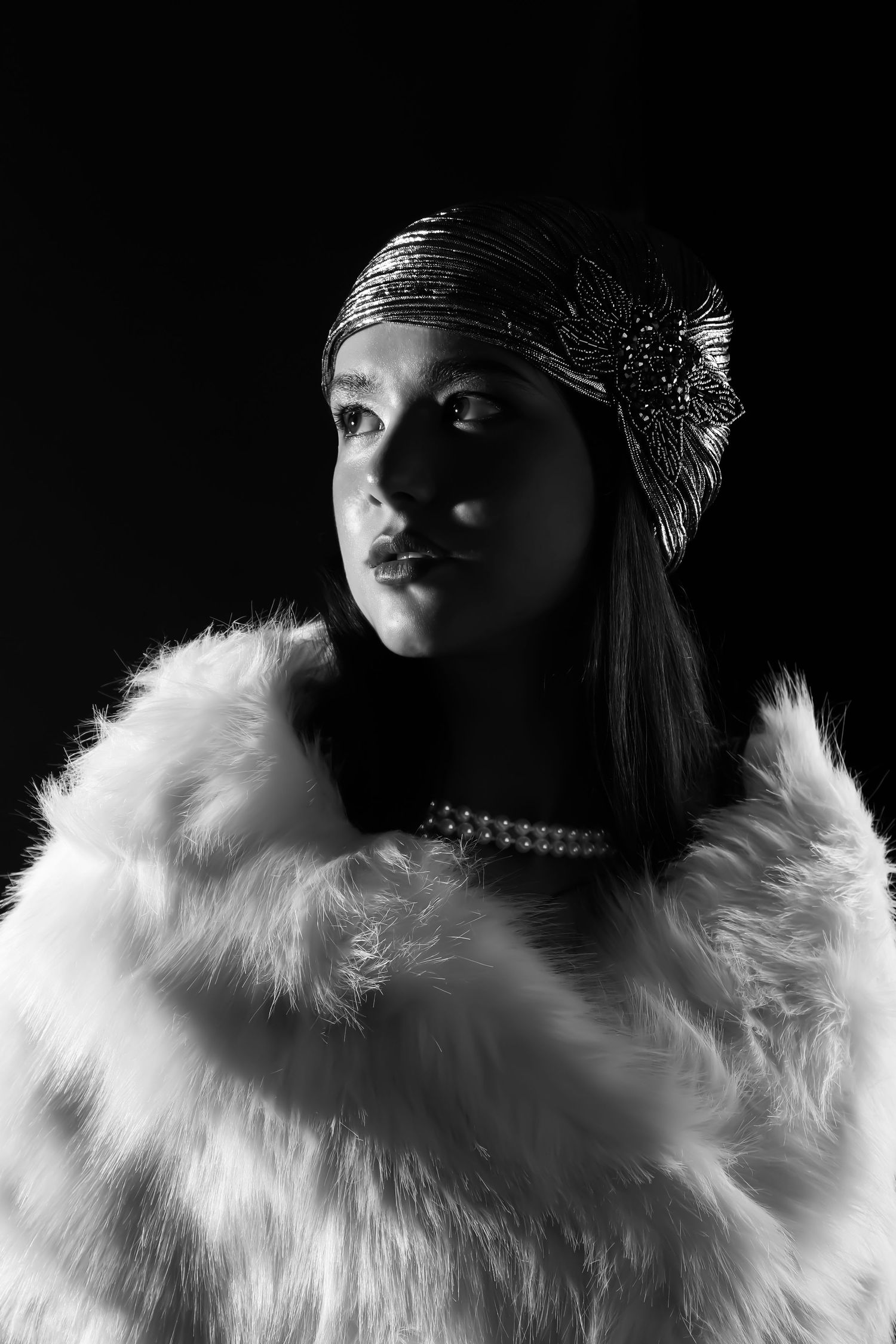 Black and white photograph of a girl' in a flapper outfit, looking up and to the left, wearing a white shall and silver hat and pearls