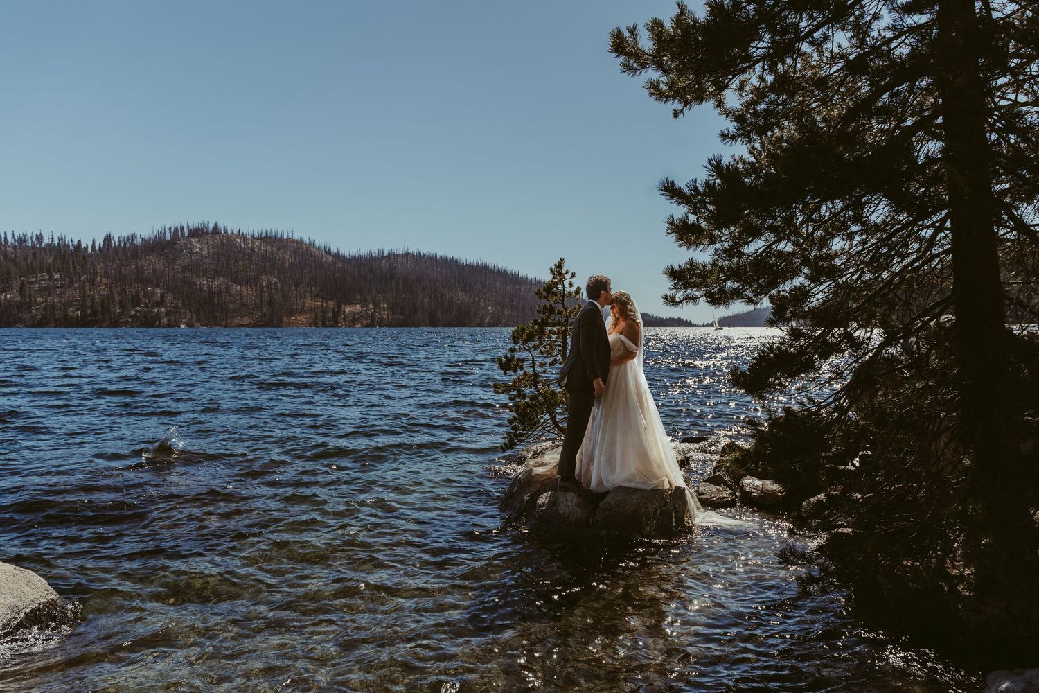 Bride and groom embracing on rocky lakeside with pine trees and mountain backdrop on a clear sunny day.