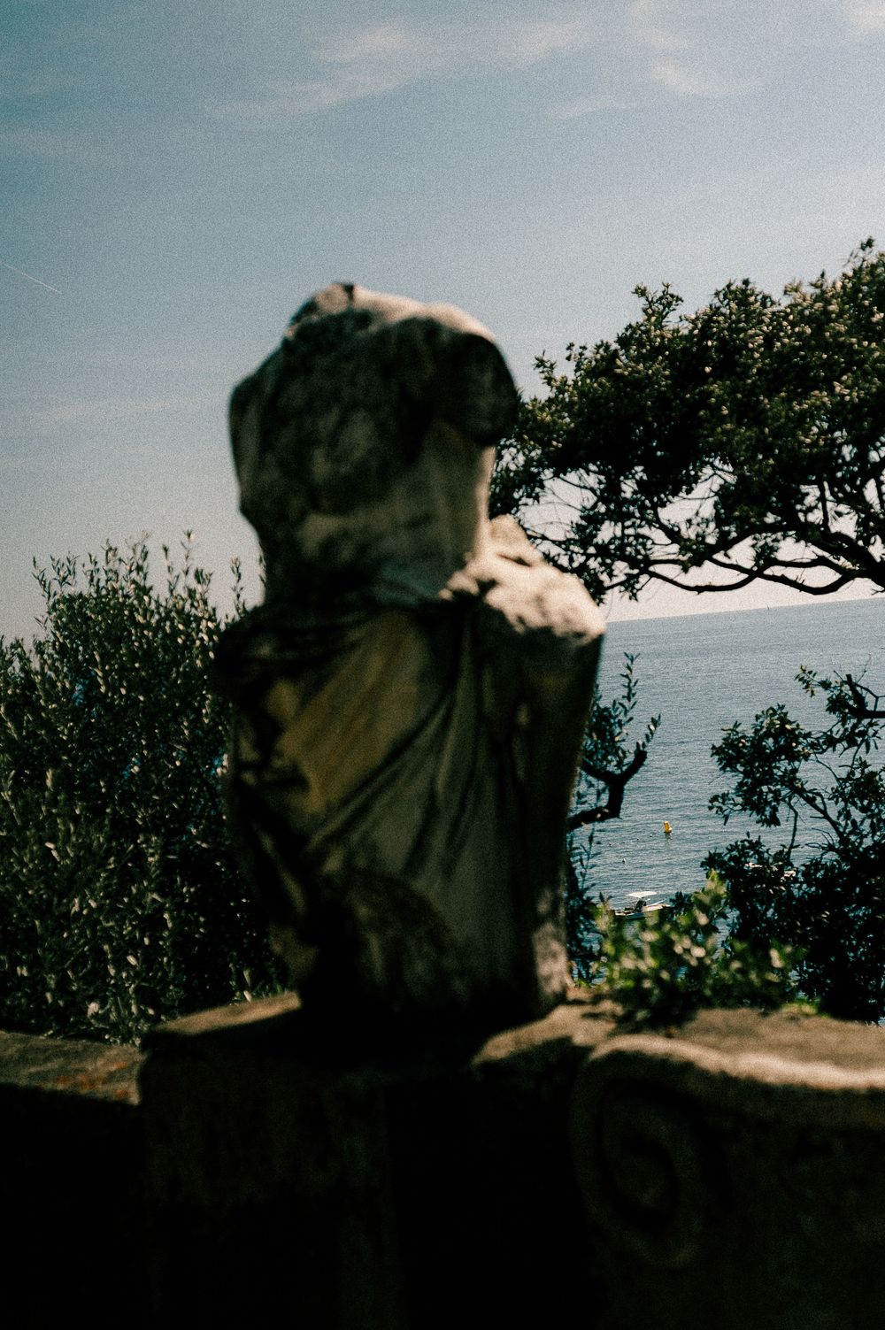 Weathered stone bust sculpture on a balustrade overlooking a serene sea, framed by lush green trees under a blue sky.