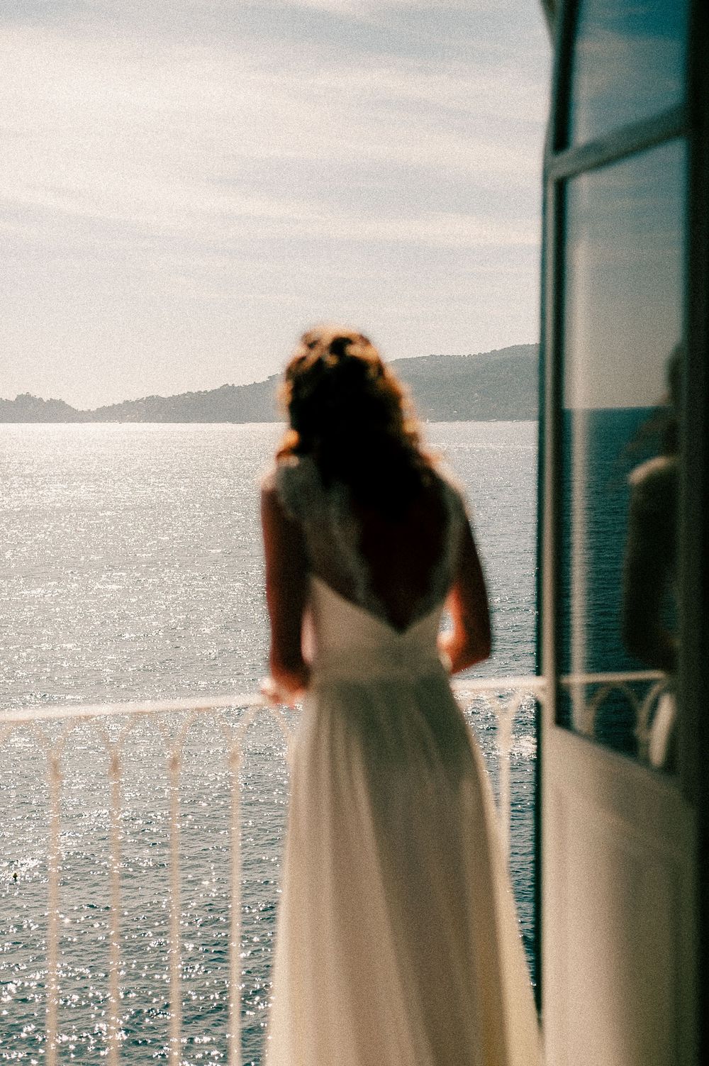 Bride in white backless gown stands on sunlit balcony overlooking sparkling ocean with distant hills.