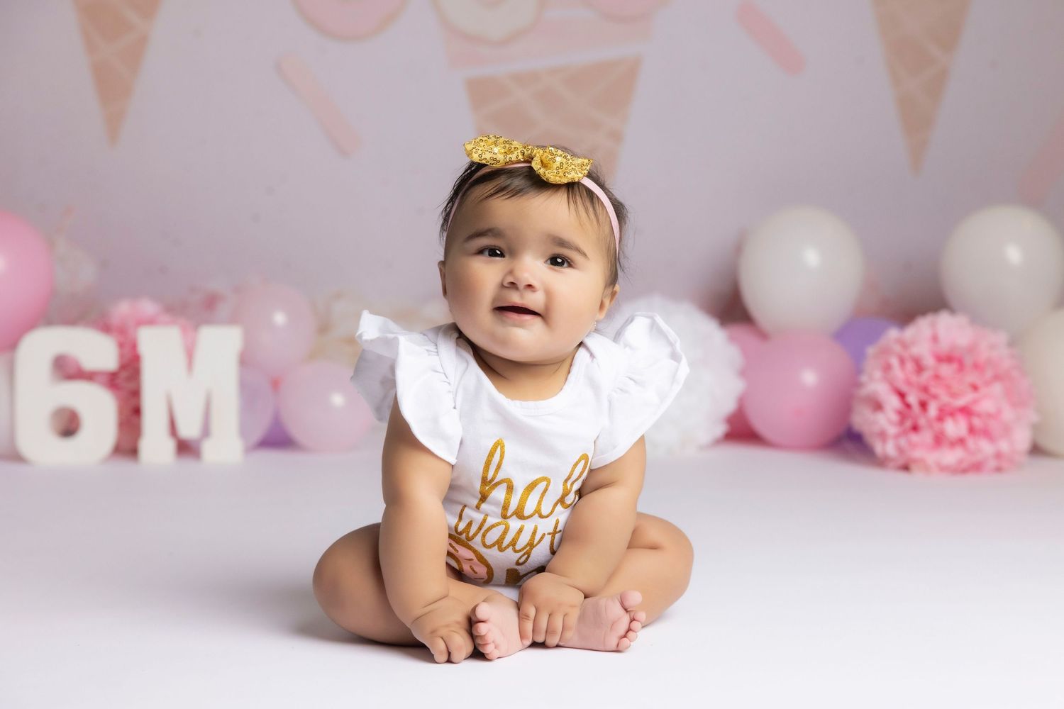 A smiling baby girl in a white romper with a gold headband sits in front of a pink balloon 6-month milestone backdrop.