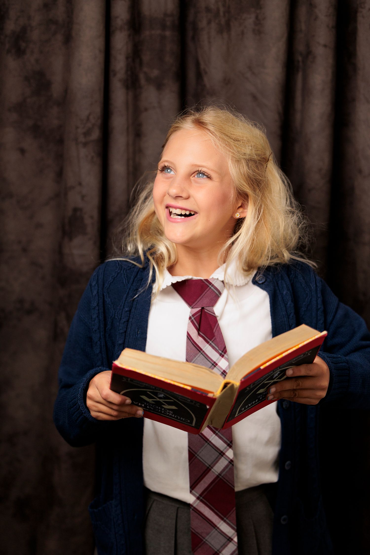 A smiling blonde girl in school uniform holds an open book while looking up with joy and excitement.