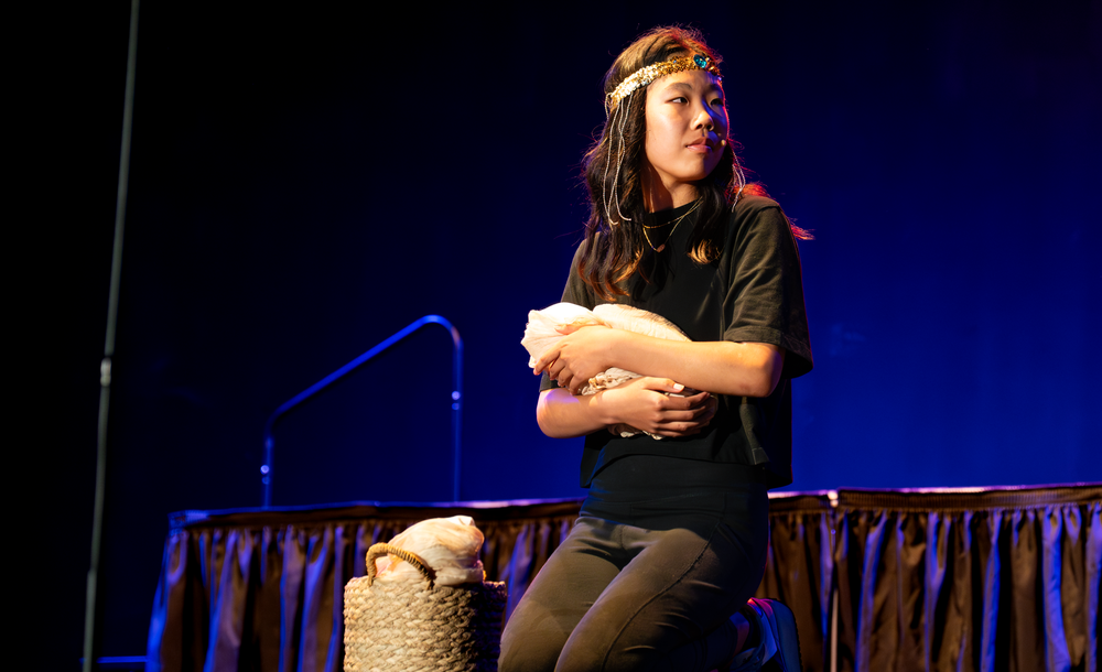 A young woman in black performs on stage, holding a white object while seated near a decorative prop.