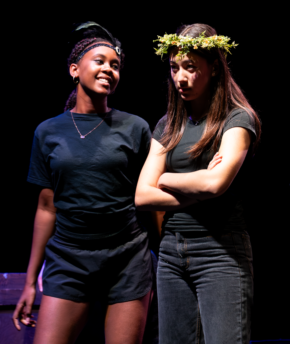 Two teenage girls in black costumes perform on stage, one wearing a floral crown with arms crossed.