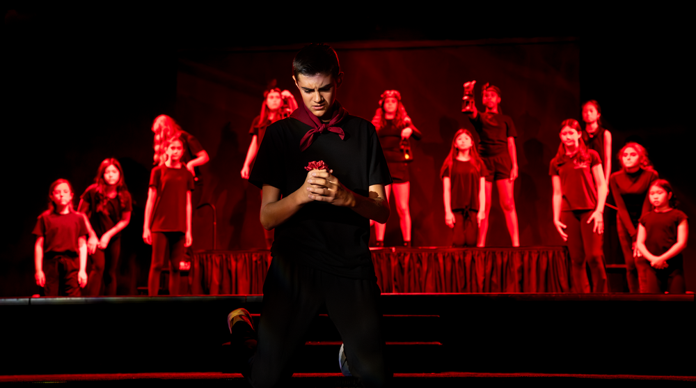 A male performer stands center stage in red lighting surrounded by a chorus of performers in red.