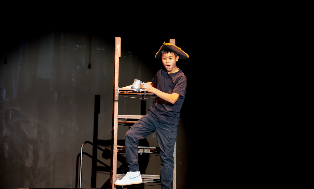 A young male actor in a hat performs a comedic scene on a ladder during a school theater production.