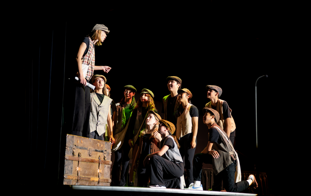 Student performers in newsboy caps gather around a wooden crate during a school stage production.