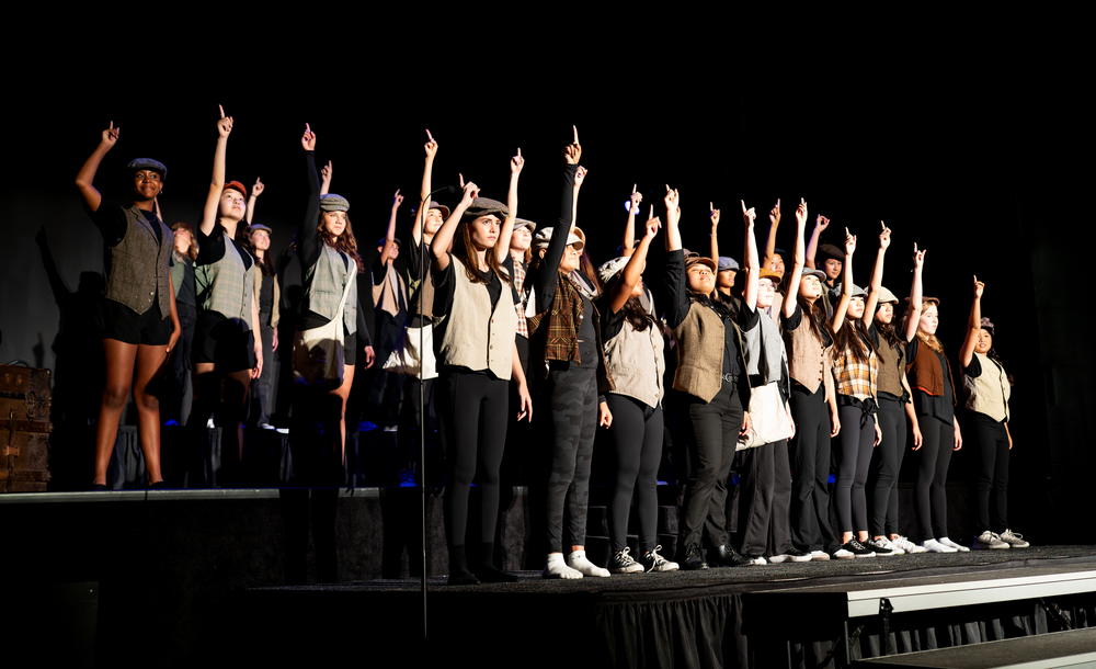 A large ensemble of student performers raises their hands in unison during a school theater finale.