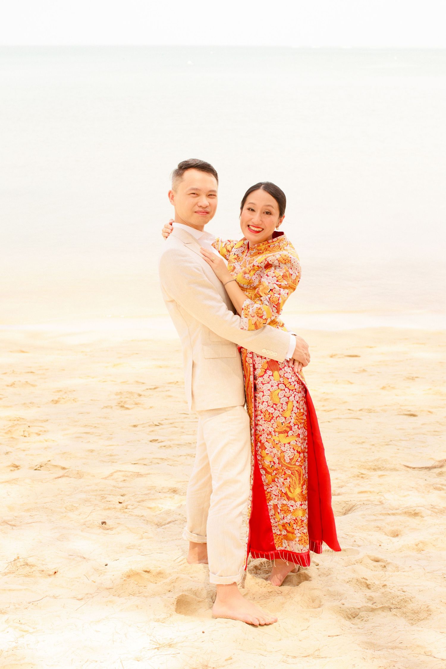 Bride in a embroided wedding cheongsam embraces groom on a sandy beach after chinese tea ceremony in Koh Samui during a destination wedding