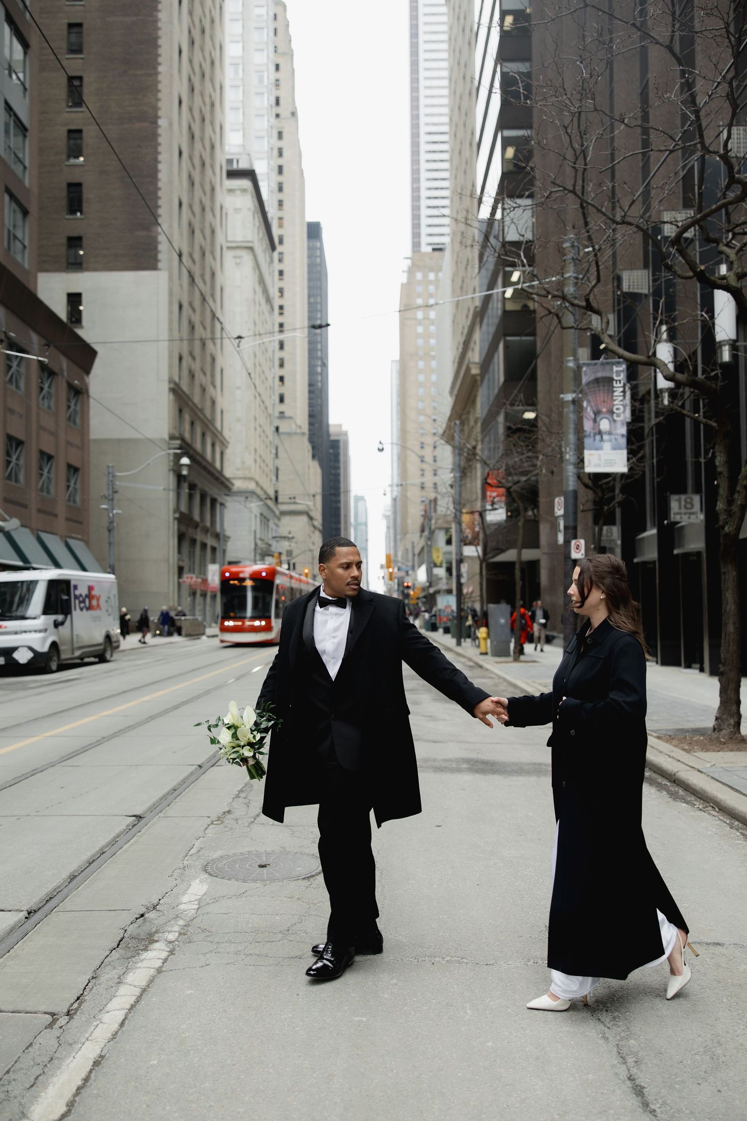 Two people in black coats perform a synchronized dance movement on an empty city street between tall buildings.