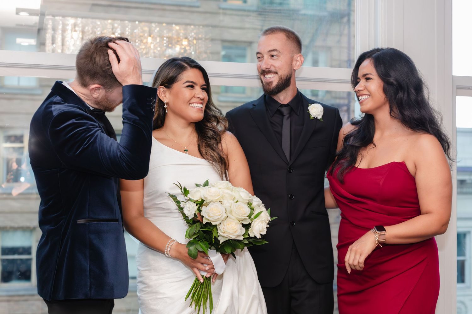 A joyful wedding party shares a candid laughing moment together on a city rooftop with the bride holding white flowers.