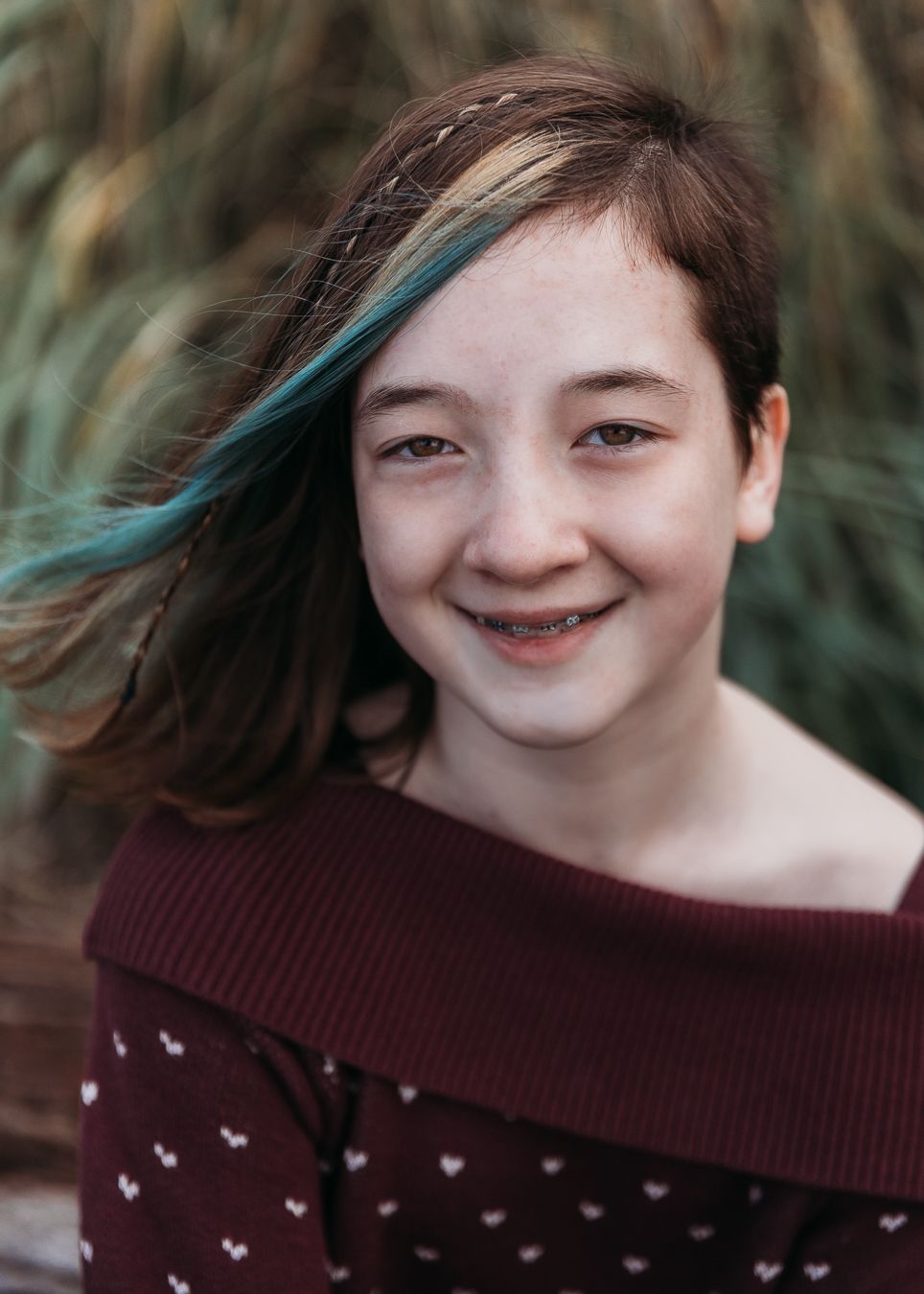 Person wearing a burgundy sweater with small white designs smiles warmly against a blurred natural background.