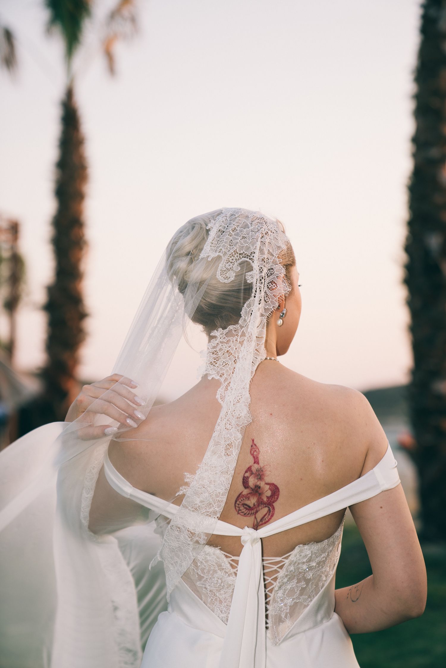 A bride in a white lace wedding dress with a veil shows her back tattoo in a series of photos at sunset.