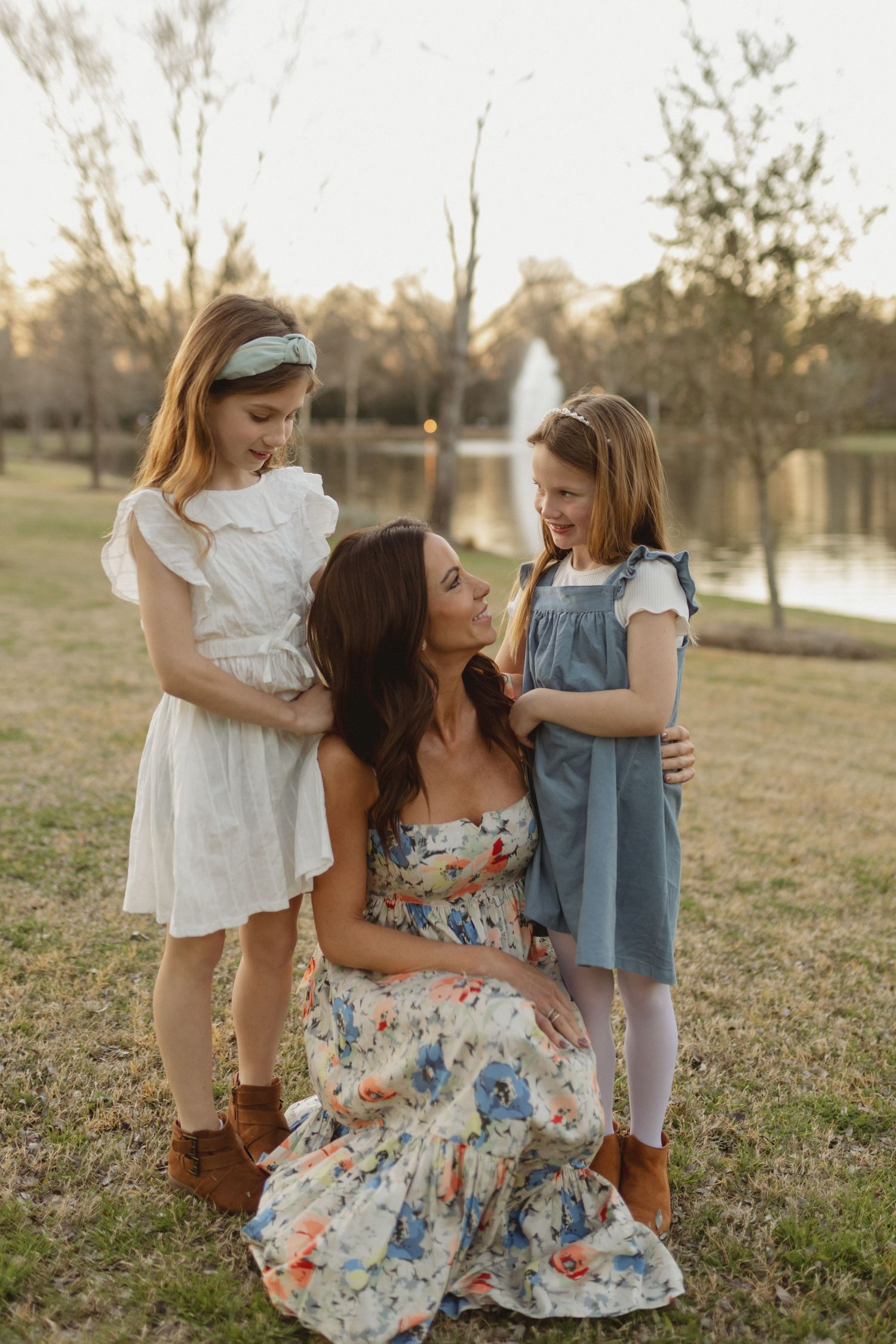 A tender family moment captured outdoors in a park with a lake and fountain in the background at sunset.