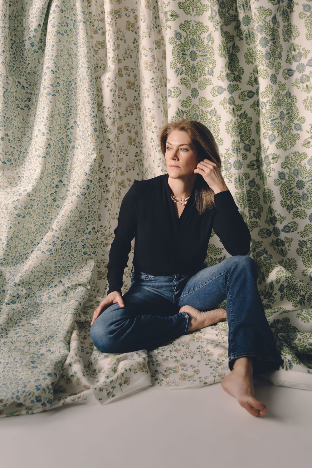 A person in a black turtleneck and jeans sits casually against a floral curtain backdrop in a minimalist setting.