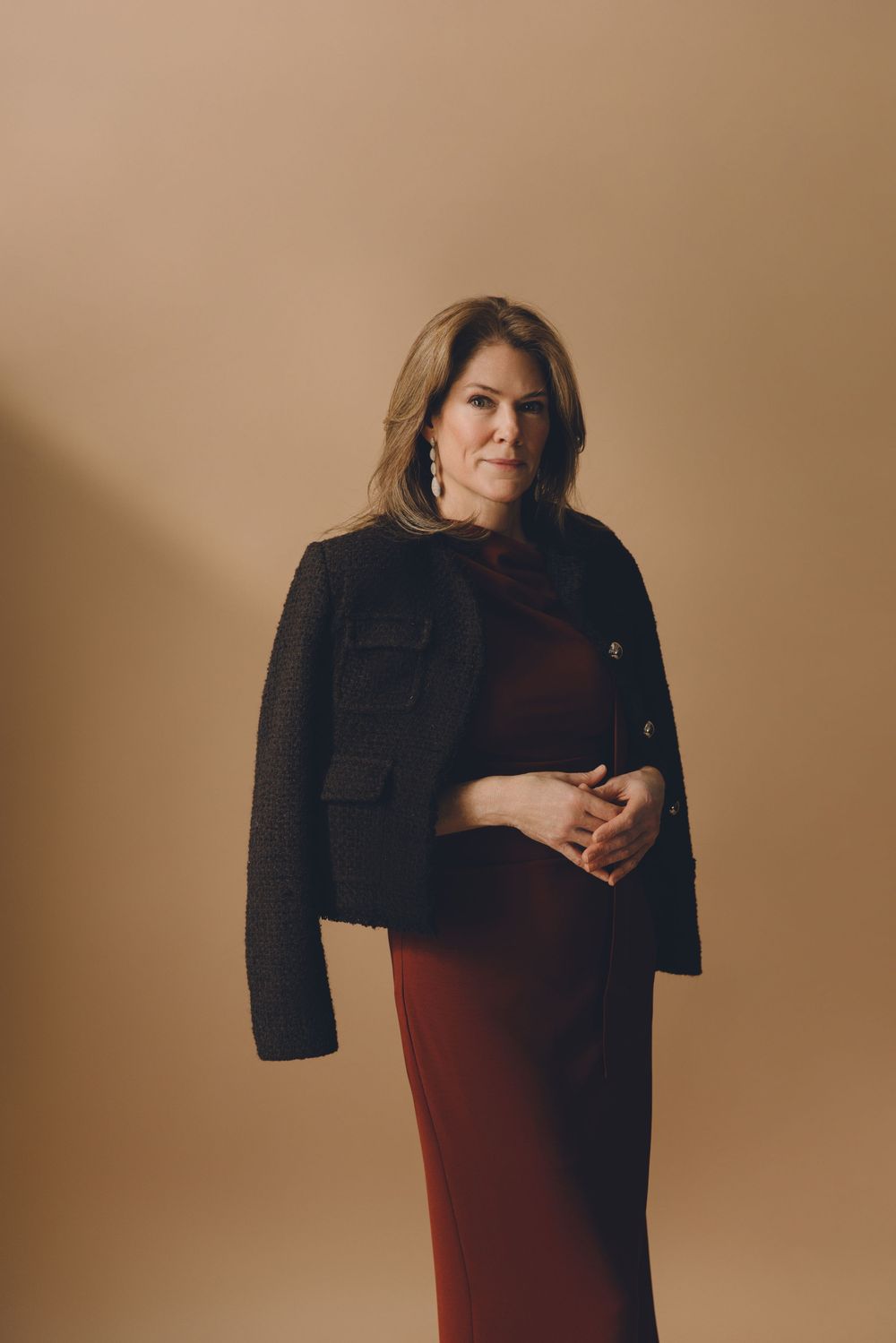 A figure in a burgundy dress holds a black blazer against a warm beige background in a minimalist fashion portrait series.