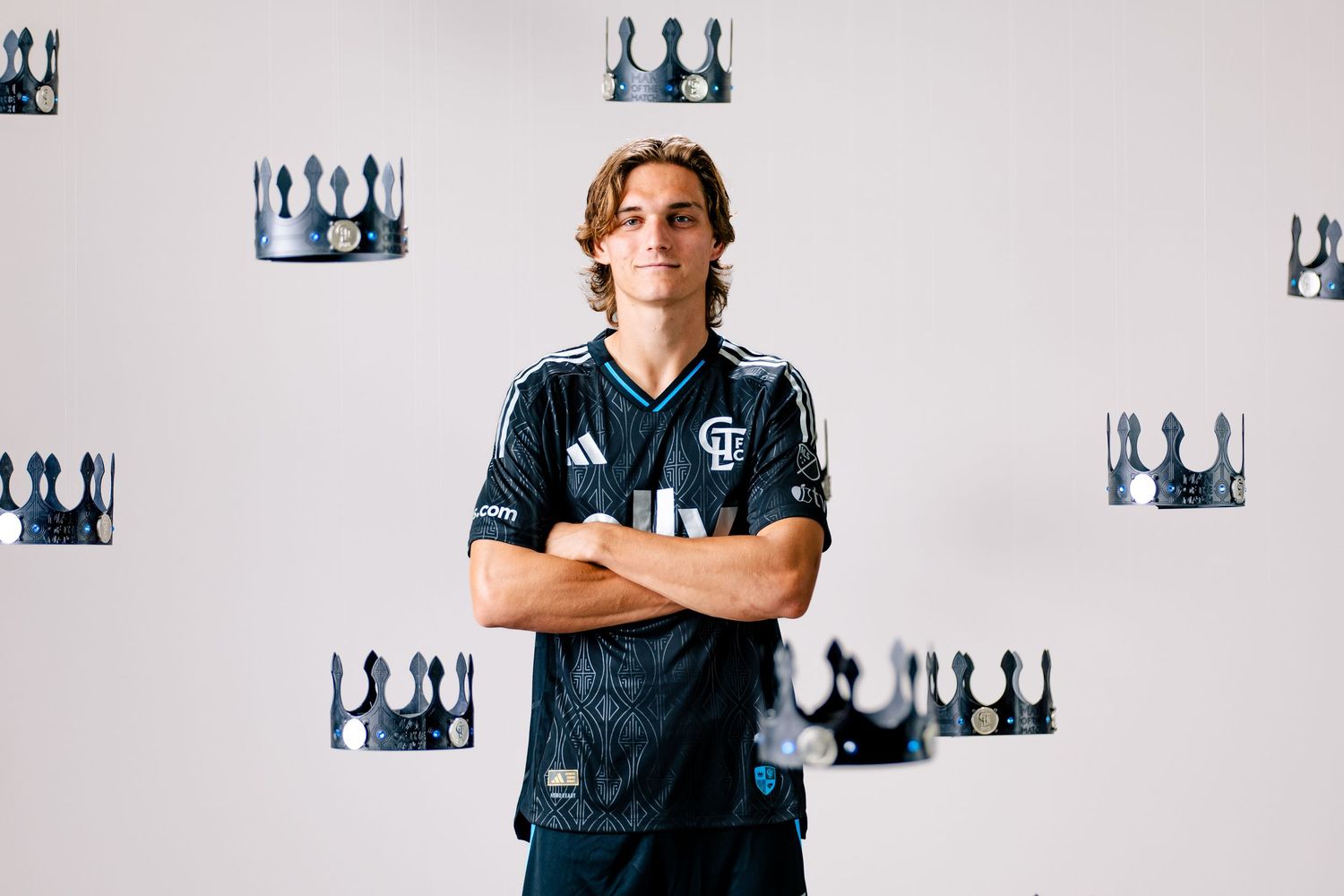 Charlotte FC player in black Adidas kit stands with arms crossed surrounded by black crown decorations on wall.