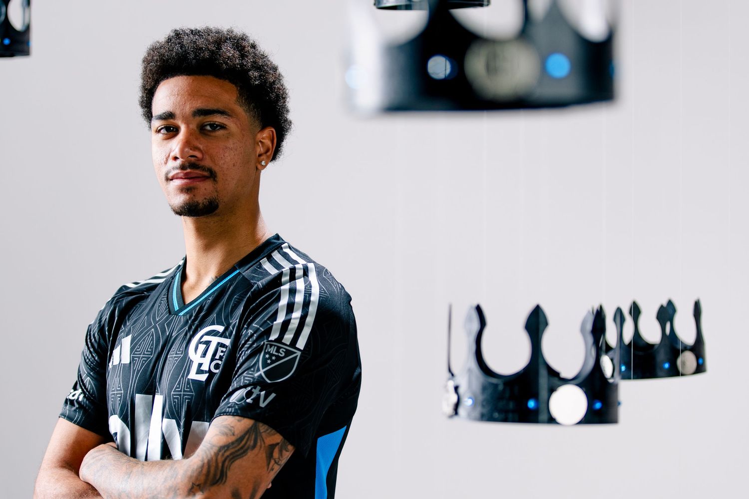 Charlotte FC player in black Adidas jersey poses with decorative black crowns in background during kit reveal.