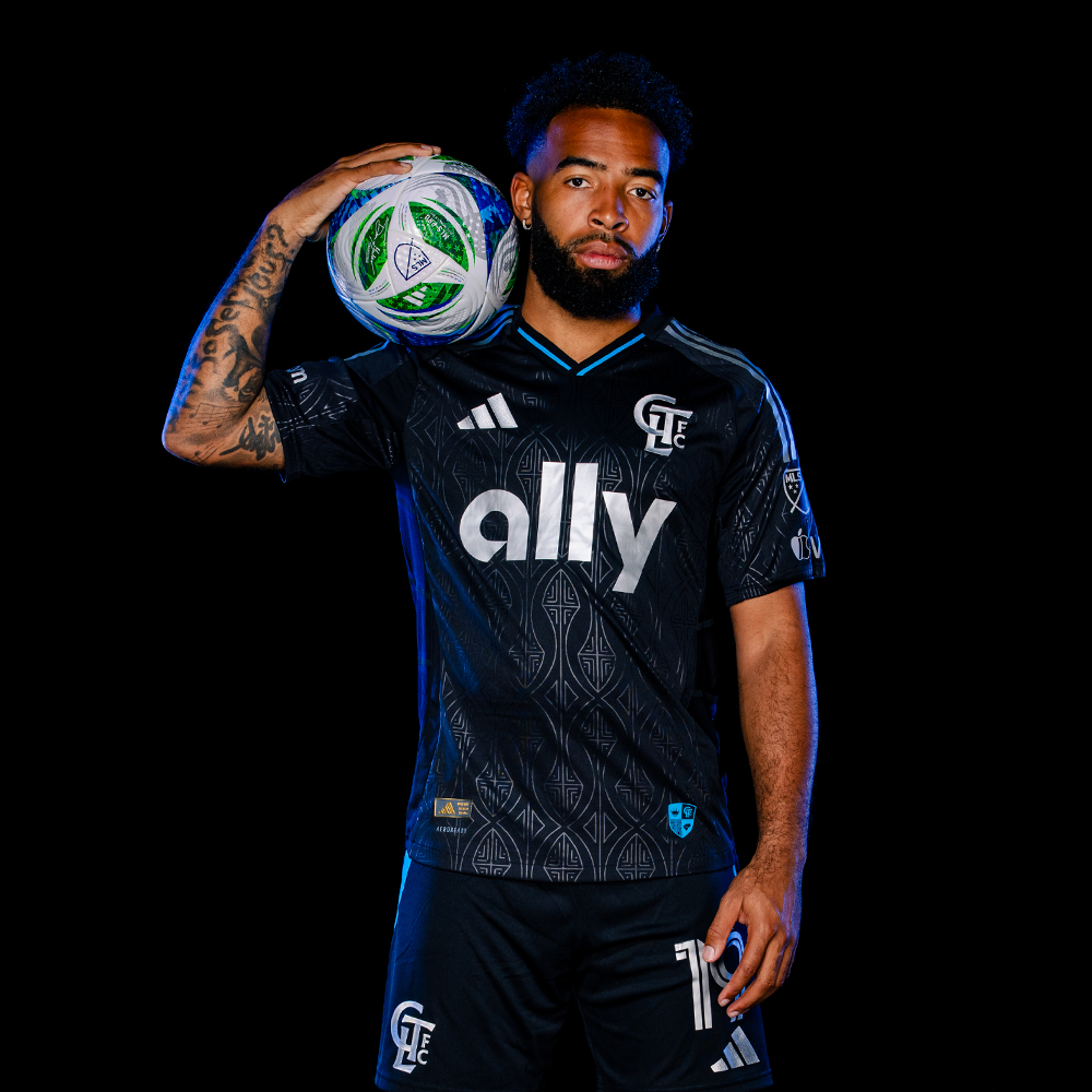 Charlotte FC player holding soccer ball on shoulder wearing black Adidas ally sponsored jersey on dark background.