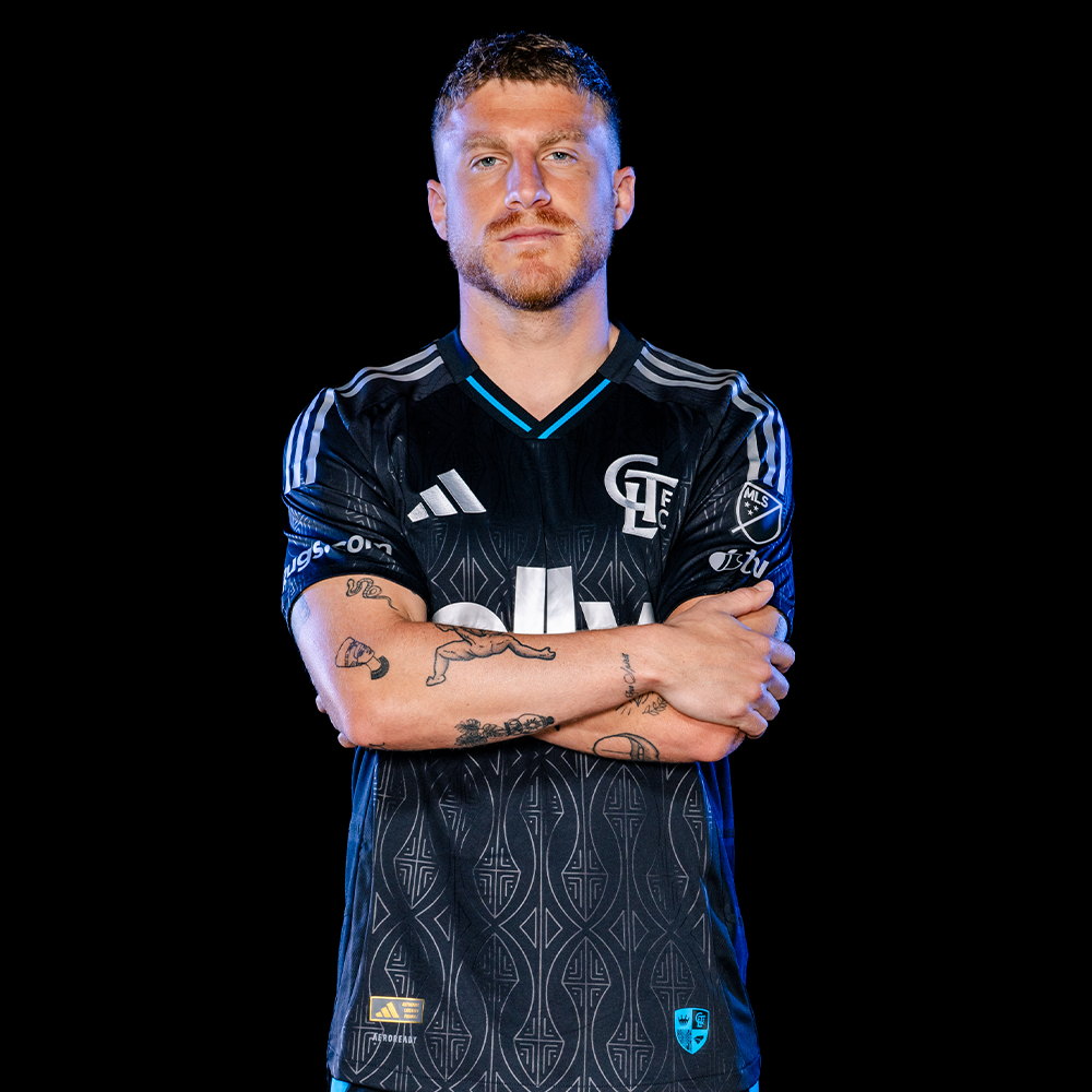 Charlotte FC player stands with arms crossed wearing black Adidas ally sponsored jersey on dark background.