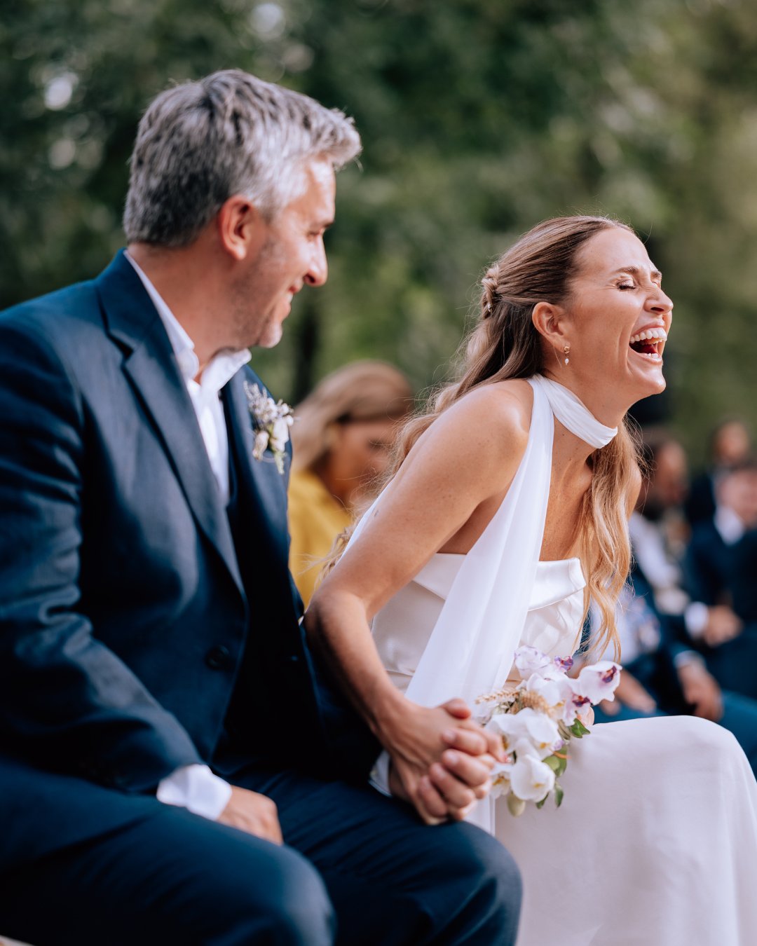 A joyful bride in white laughs beside her groom in a navy suit during an outdoor wedding ceremony.