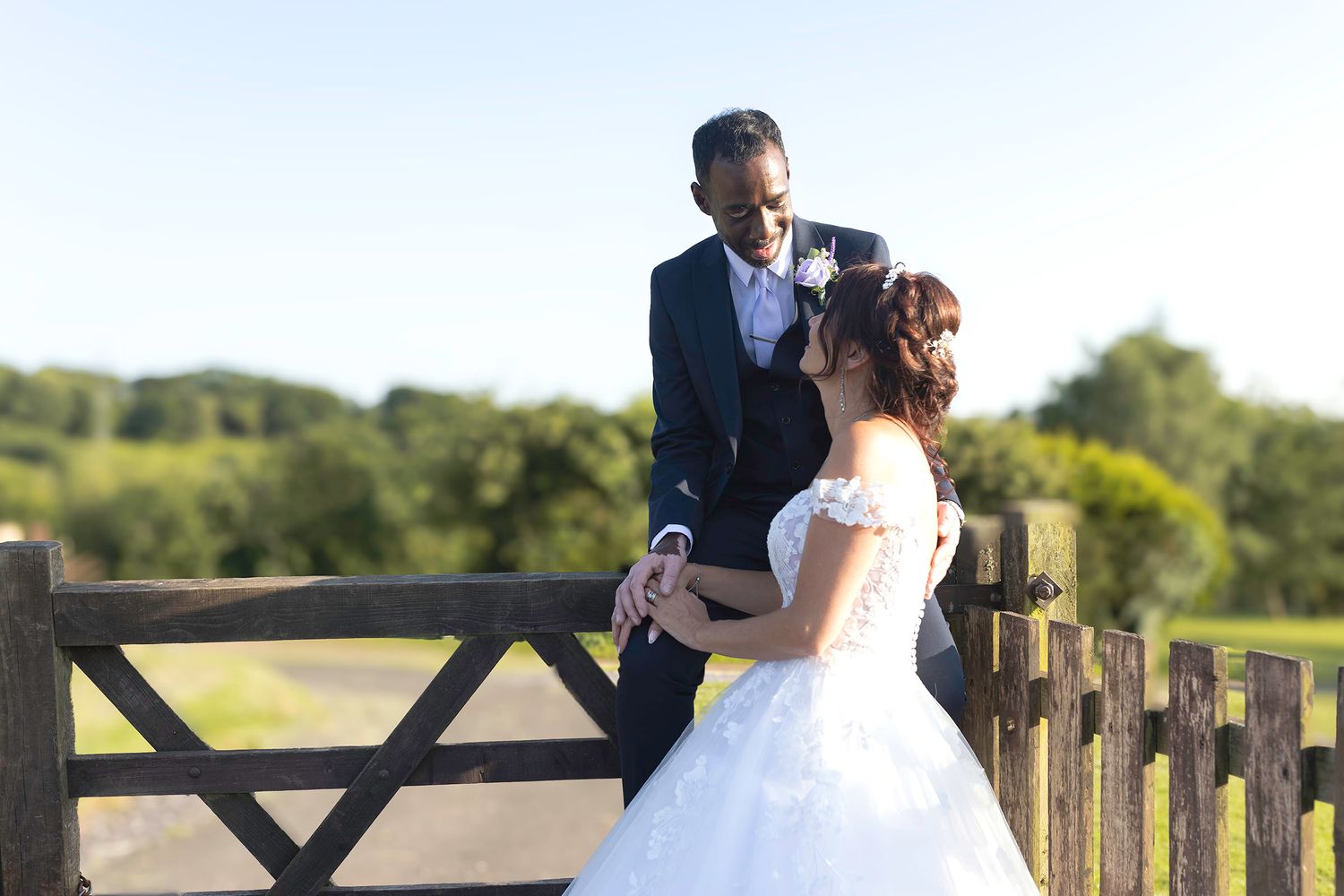 Bride and groom holding hands by a rustic wooden fence with scenic green countryside in golden sunlight.